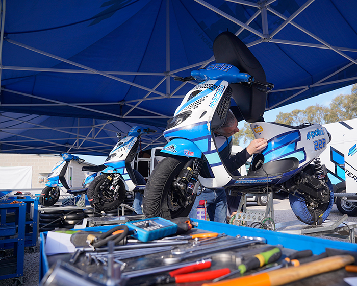 Meccanico che lavora su scooter da corsa Polini con attrezzi in primo piano e un altro scooter Polini sullo sfondo. Preparazione gara scooter.